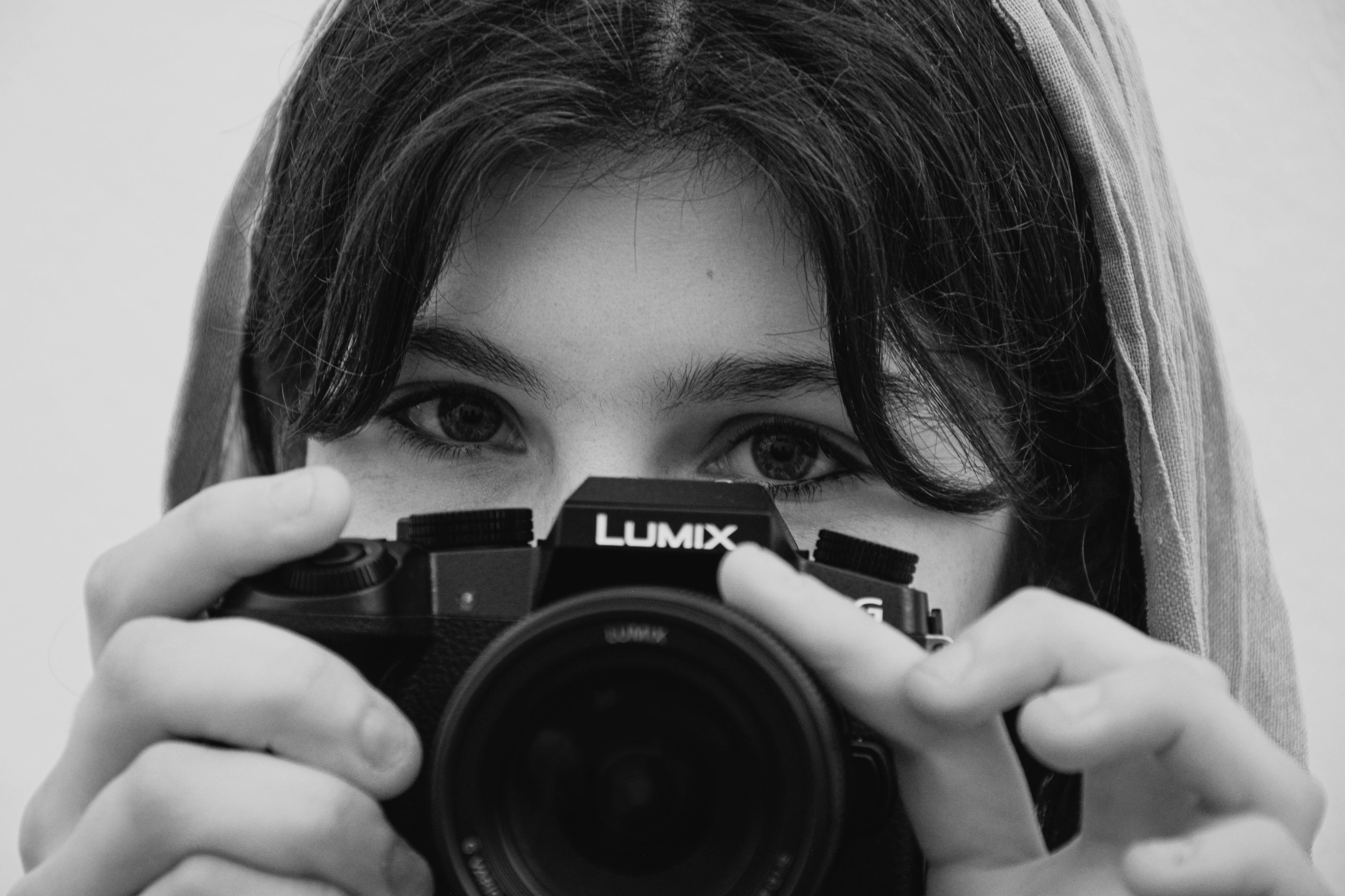 Black and white portrait of a girl holding a Lumix camera, making eye contact.