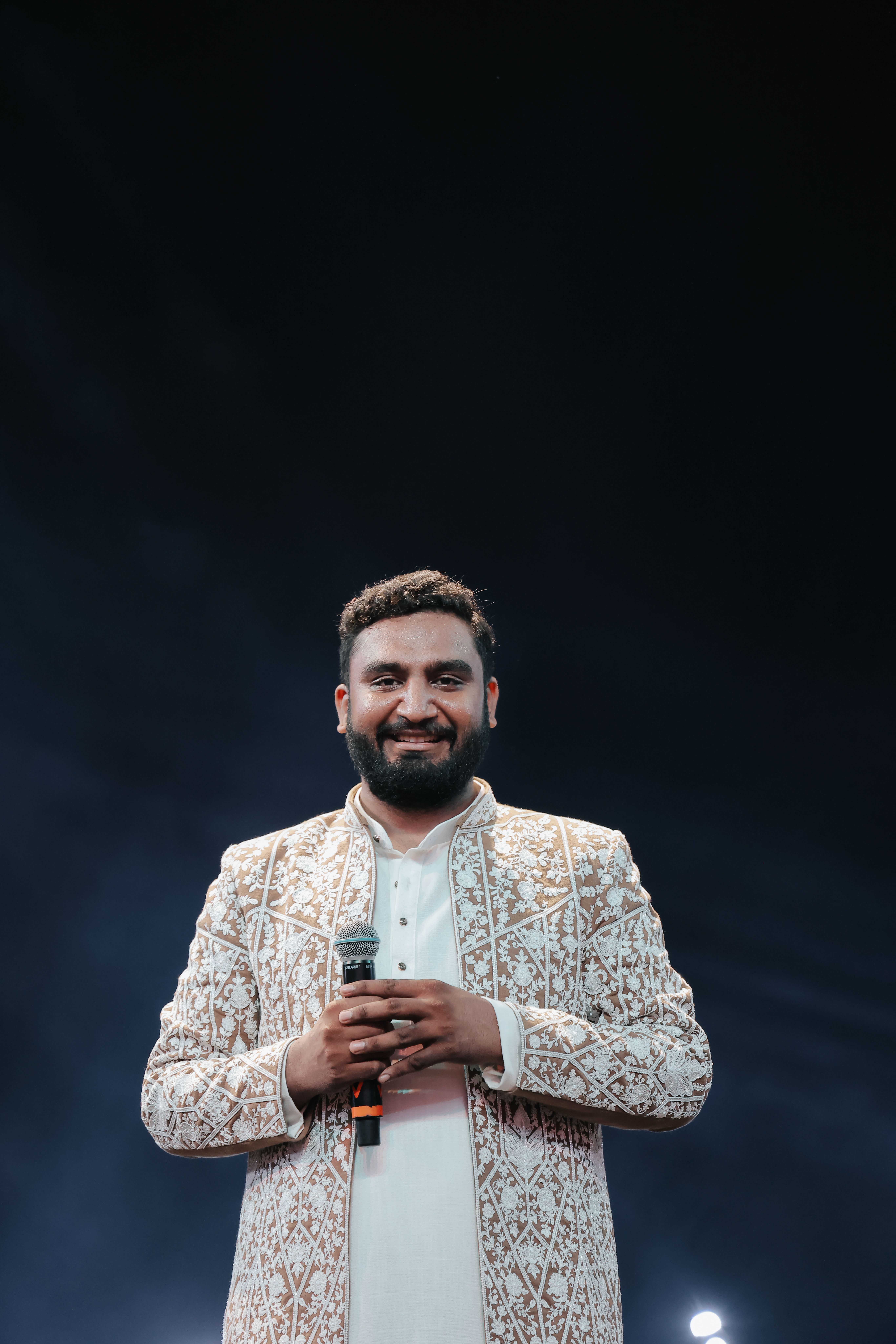 Captivating portrait of an adult man with traditional embroidered attire holding a microphone, smiling on a stage.