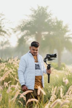 A photographer in casual attire holding a gimbal camera amidst a grassy field, captured during daylight.