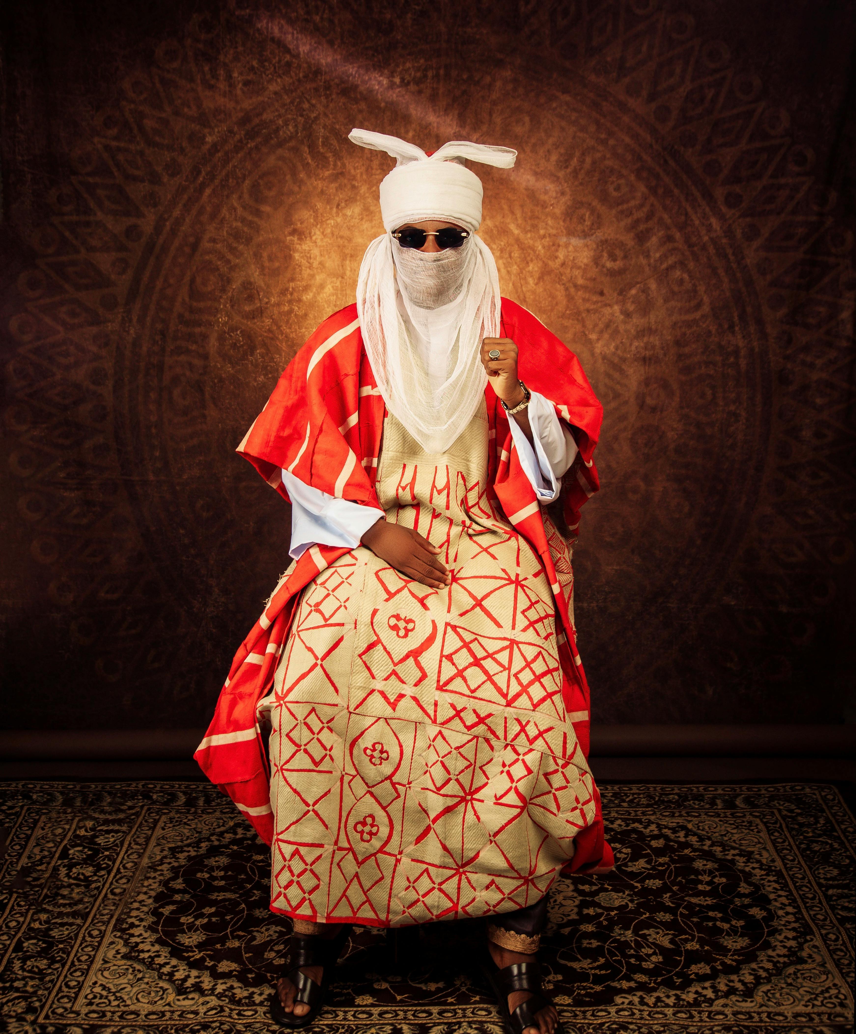 Portrait of a person in traditional African attire seated against ornate backdrop.