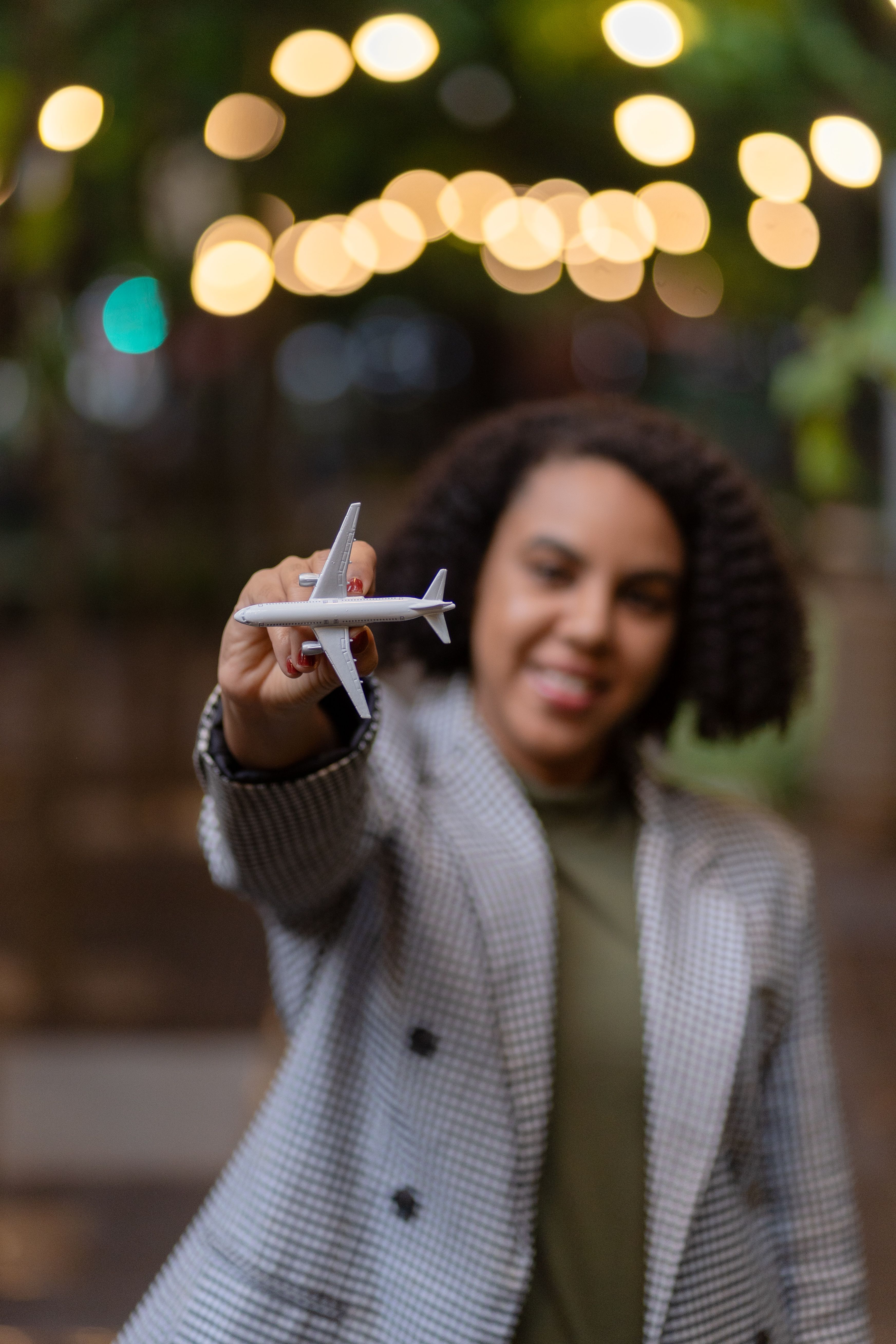 A young woman in a checkered coat holding a toy airplane with city lights in the background.