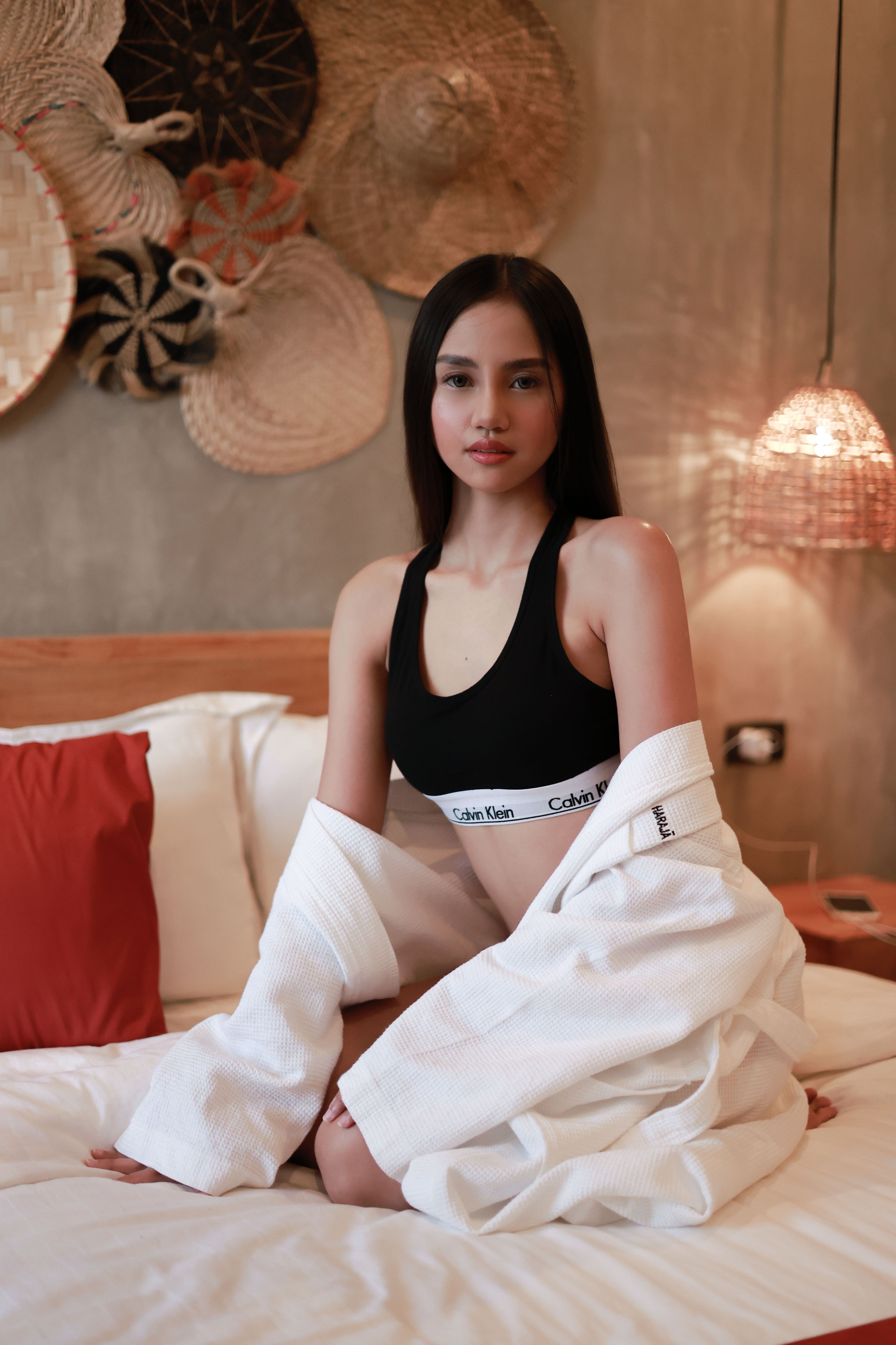 Young woman in casual wear poses confidently on bed with stylish decor in the background.
