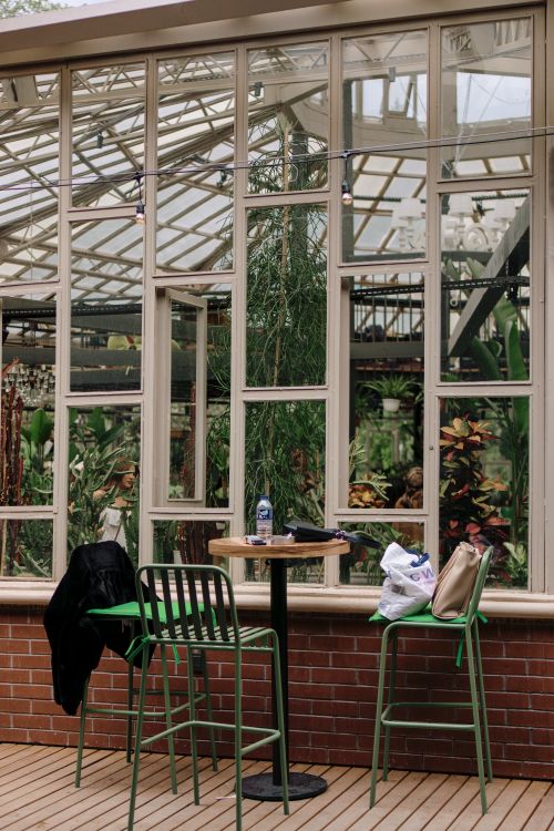 Free Peaceful greenhouse cafe scene with empty chairs and lush plants visible through glass windows. Stock Photo
