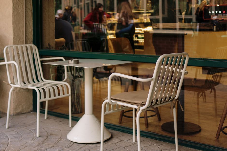 Charming outdoor cafe seating with white chairs and a table beside a vintage window reflecting a warm ambiance.