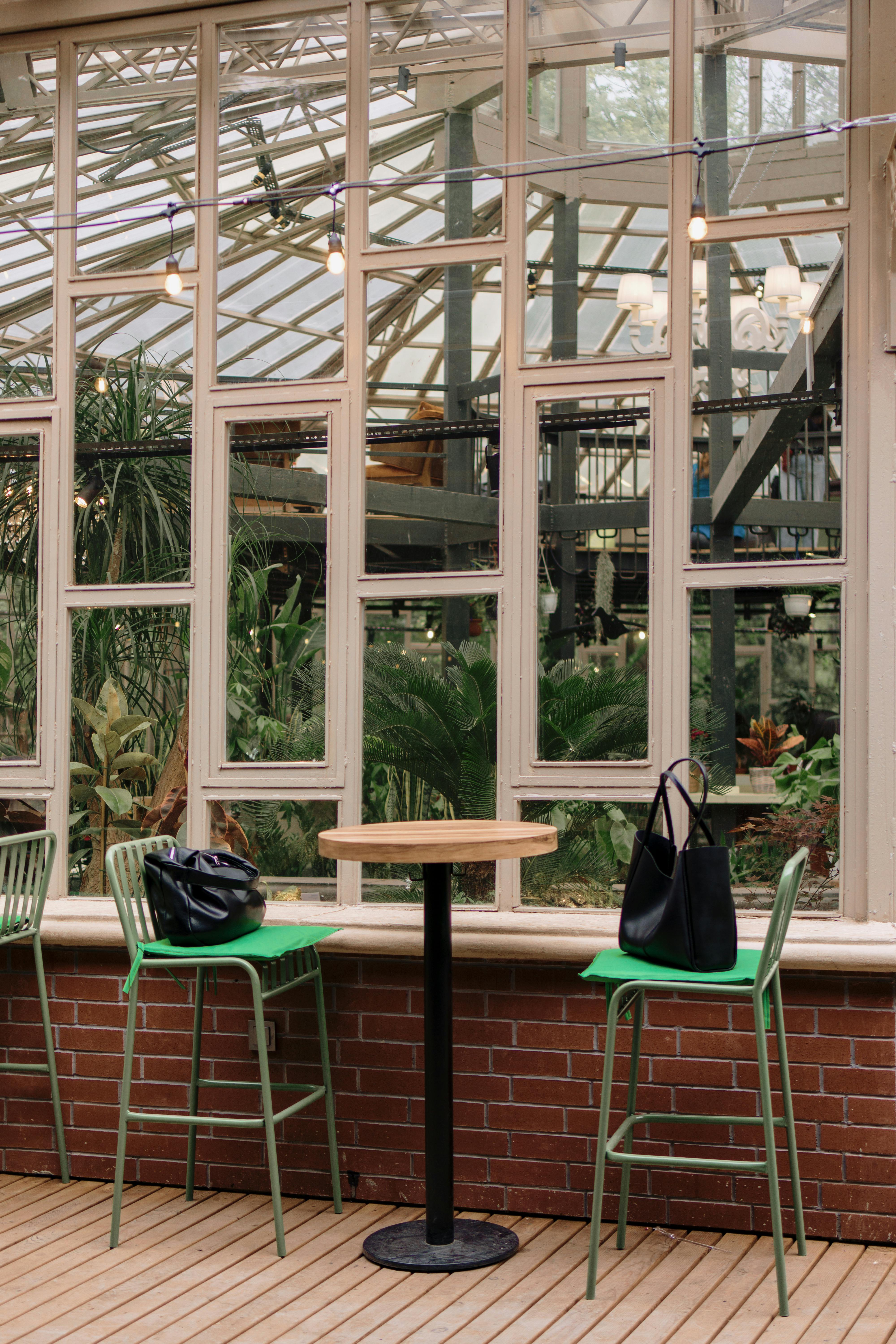 Cozy indoor cafe with a view of a lush greenhouse, featuring green chairs and a central table.