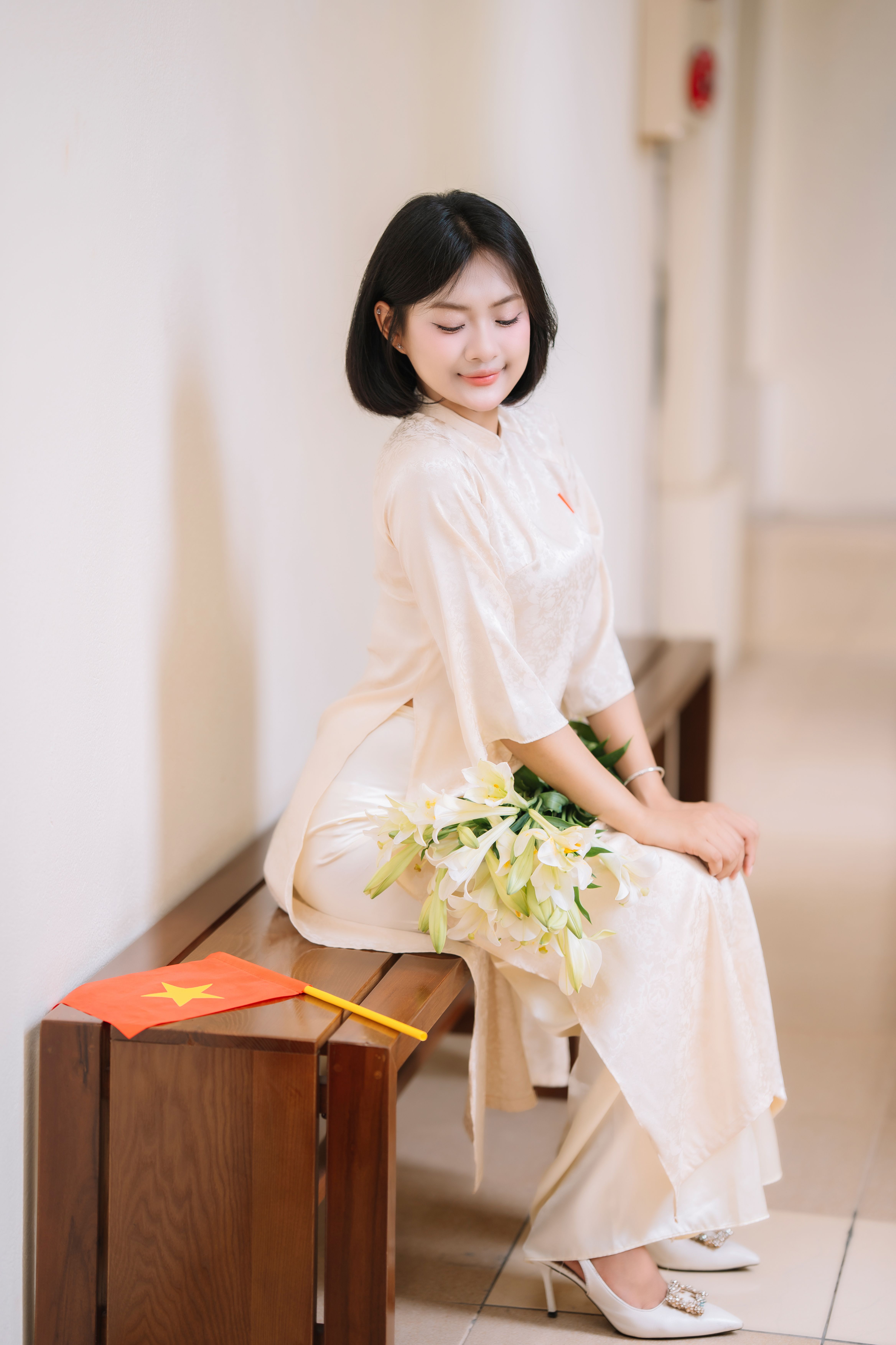 A young woman seated indoors in an elegant Vietnamese dress with a bouquet and a Vietnamese flag.