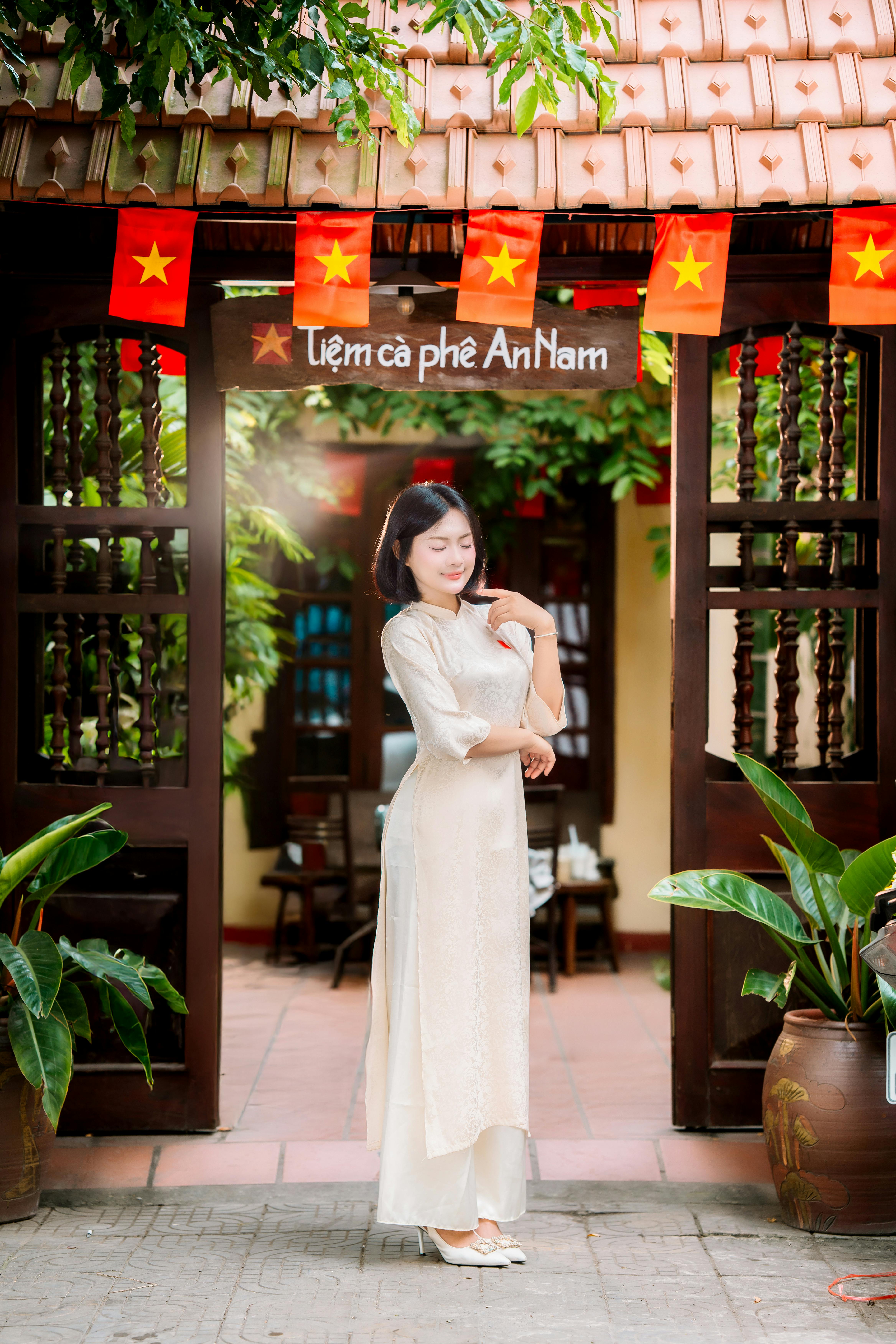 A woman in a traditional Ao Dai poses gracefully at a Vietnamese café adorned with flags.