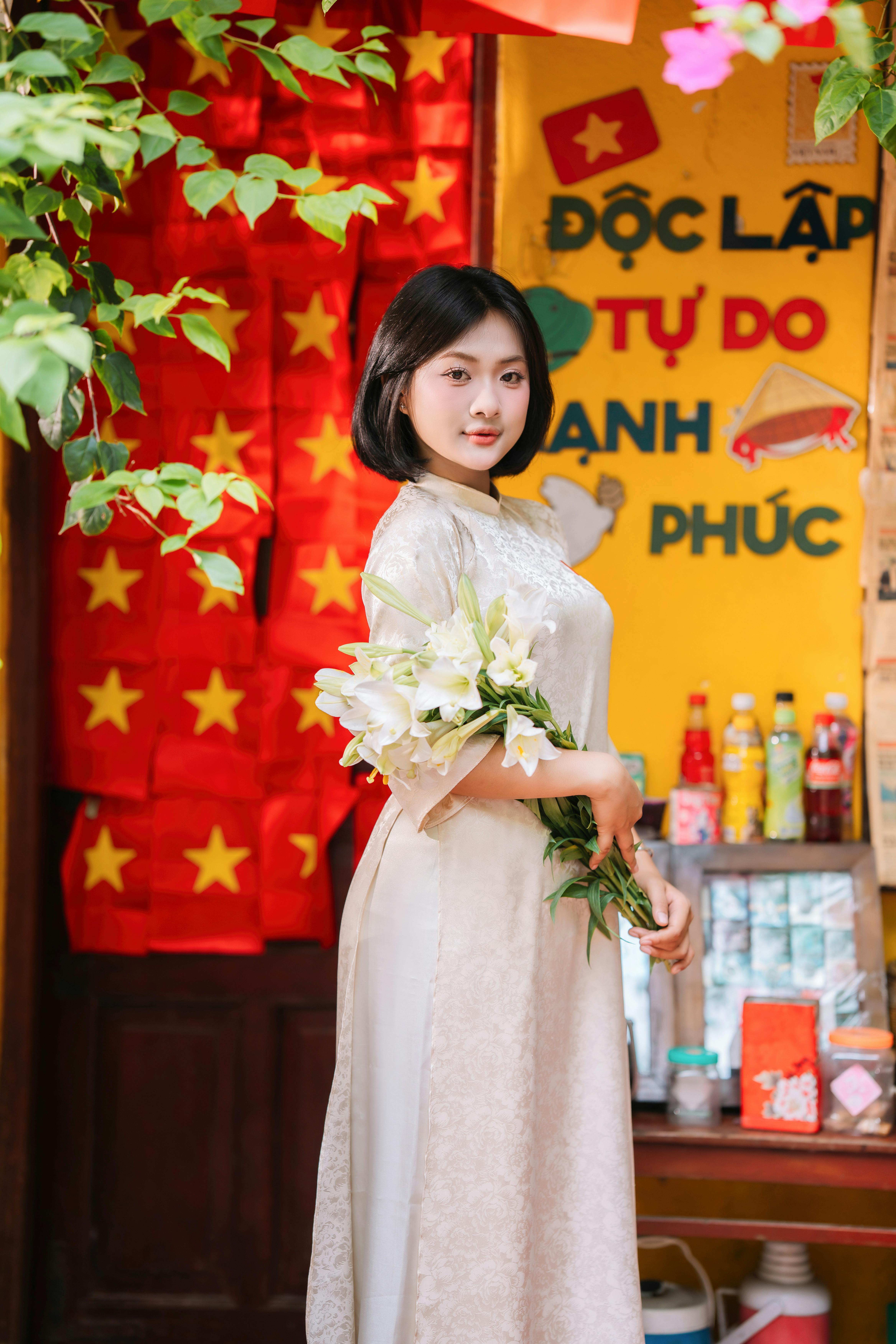 Young Vietnamese woman in traditional dress holding flowers, stands by national flags.