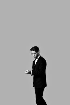 Black and white portrait of a stylish man in formal attire checking his phone against a minimalist background.