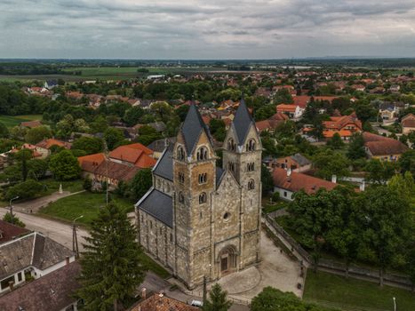 Aerial view of a Romanesque church surrounded by lush greenery and red-roofed houses in a Hungarian village.
