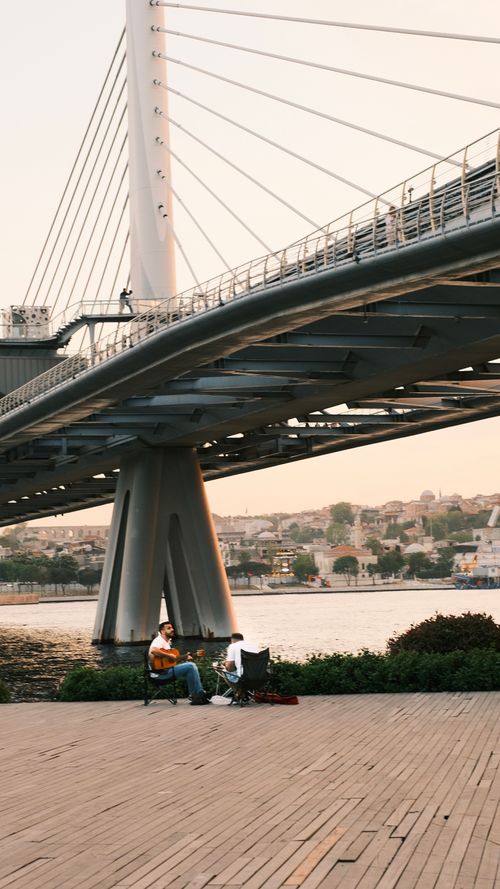 Free A tranquil scene of musicians playing guitar beneath a modern bridge at sunset. Stock Photo