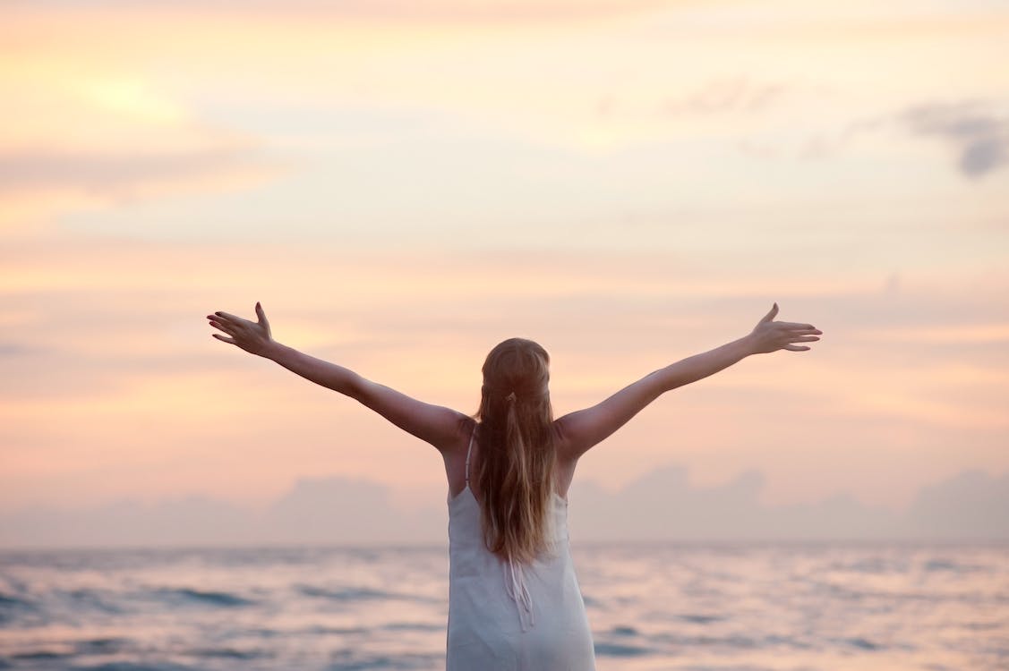 Free Rear View of Woman With Arms Raised at Beach during Sunset Stock Photo