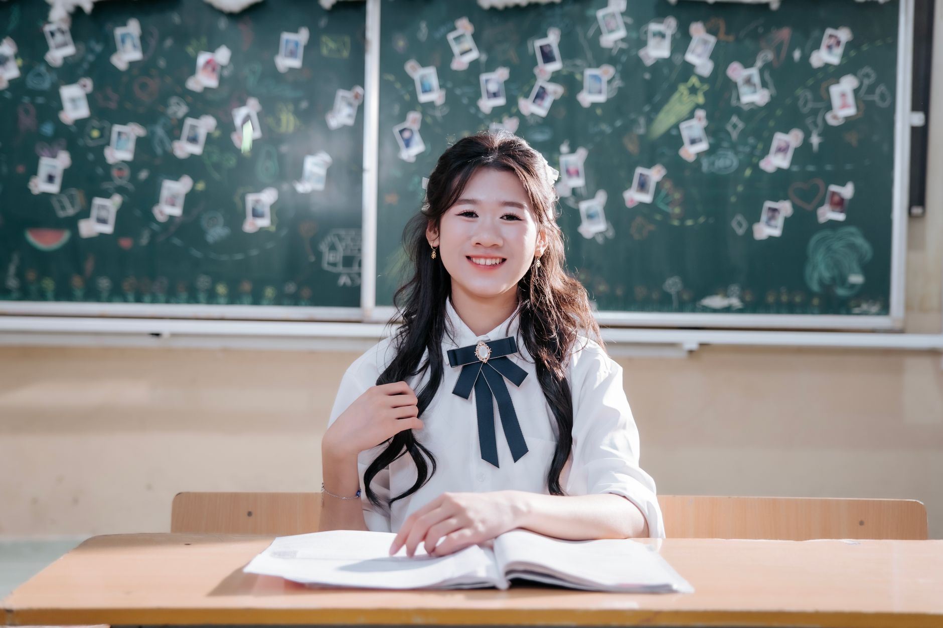 A cheerful student sitting at a desk in a classroom with decorated chalkboard.