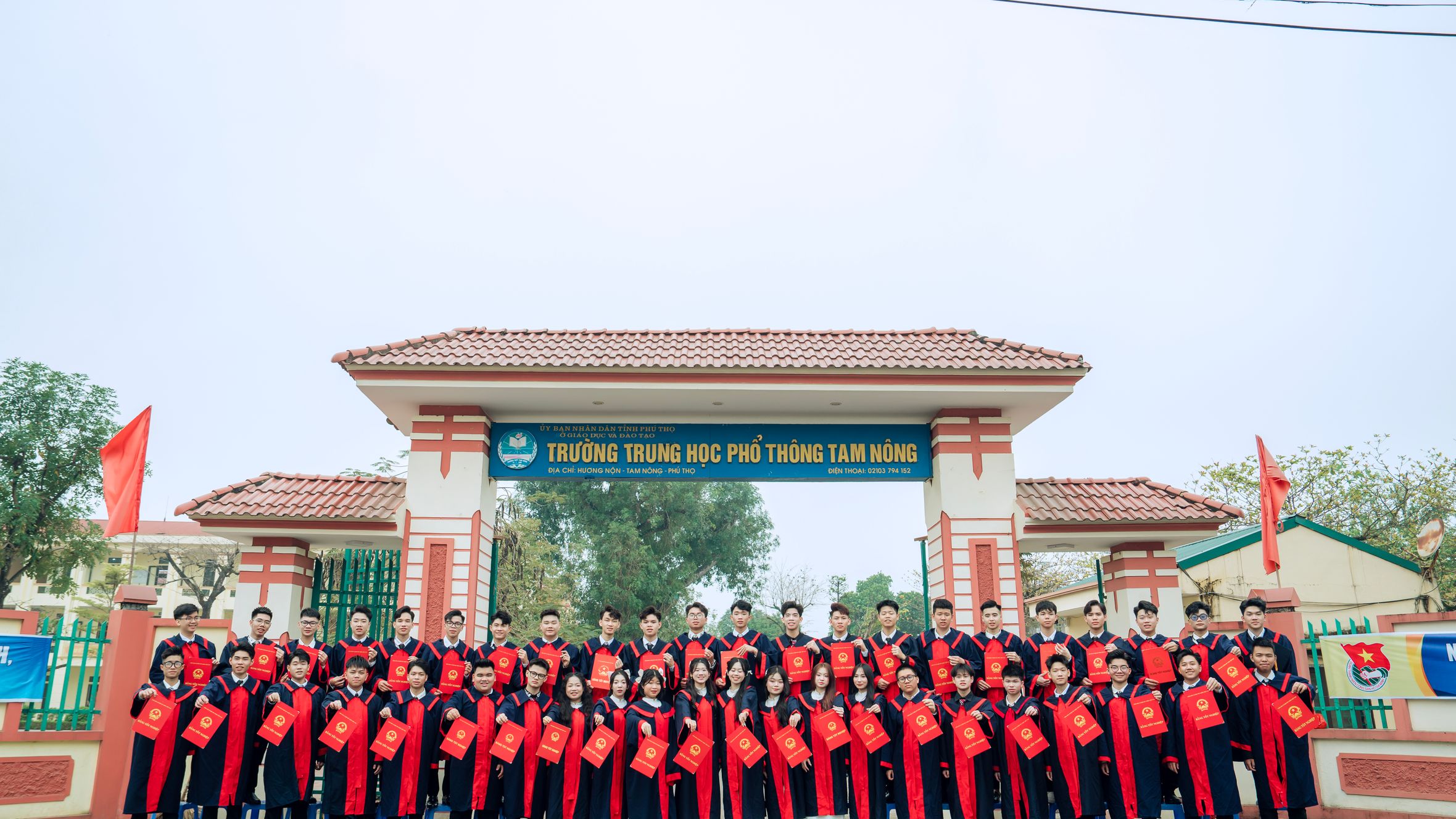 Large group of students in graduation robes posing in front of Tam Nong High School gate.