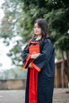 Portrait of a young adult female graduate in traditional cap and gown holding a diploma outdoors.