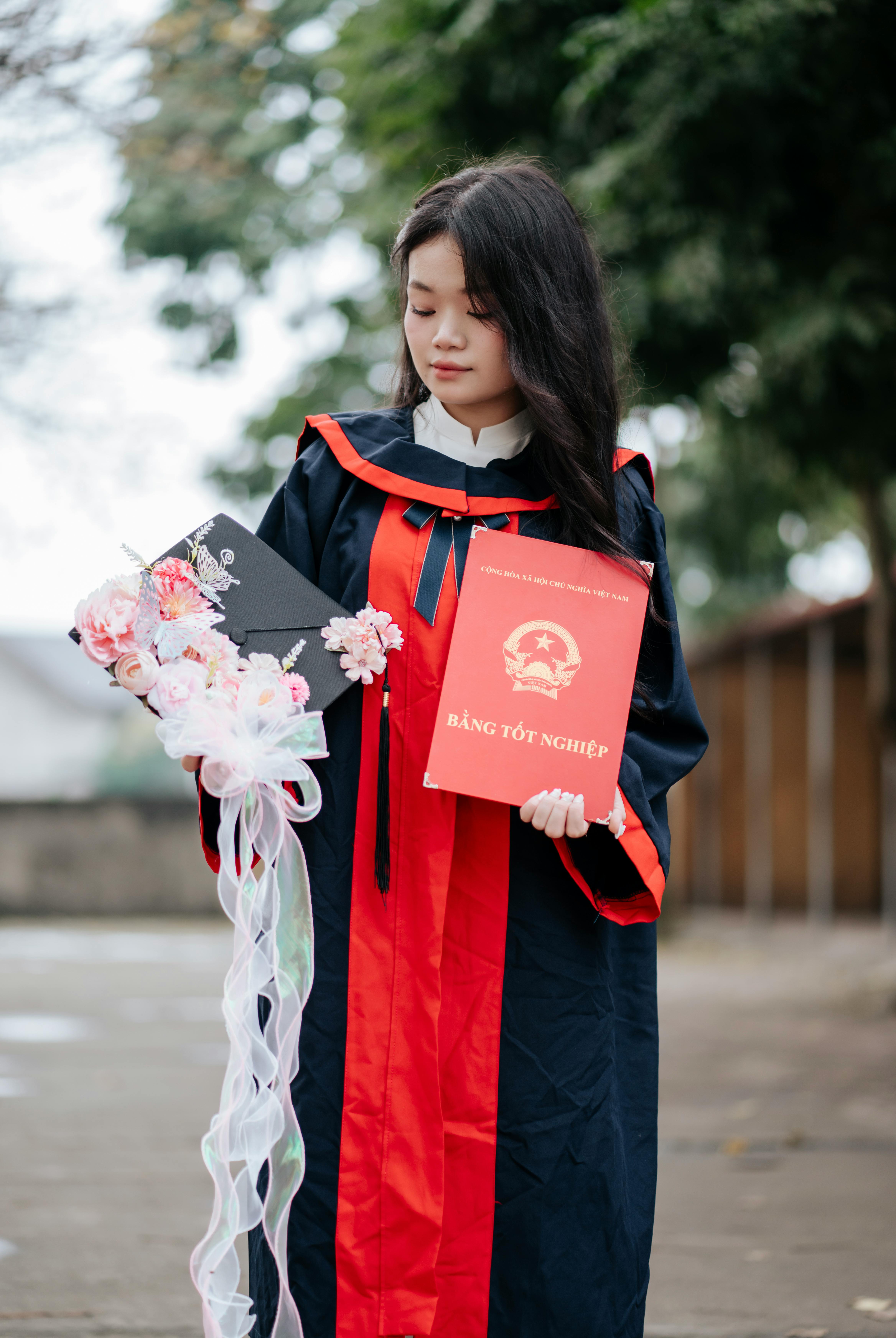 Young Asian woman in graduation attire holding diploma and cap with floral decoration outdoors.