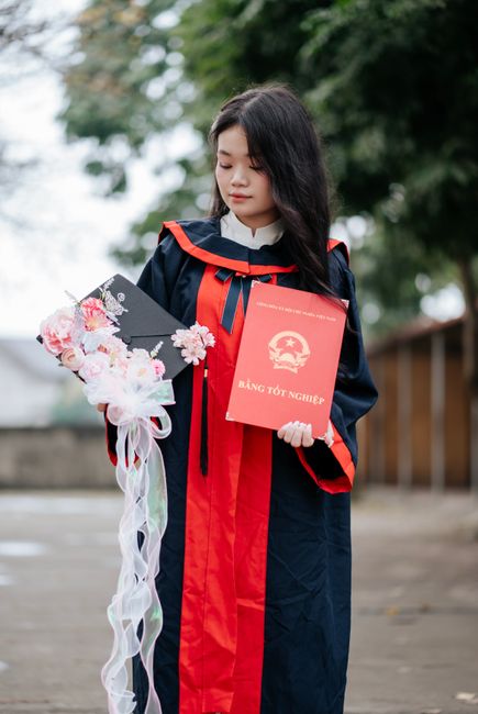 Young Asian woman in graduation attire holding diploma and cap with floral decoration outdoors.