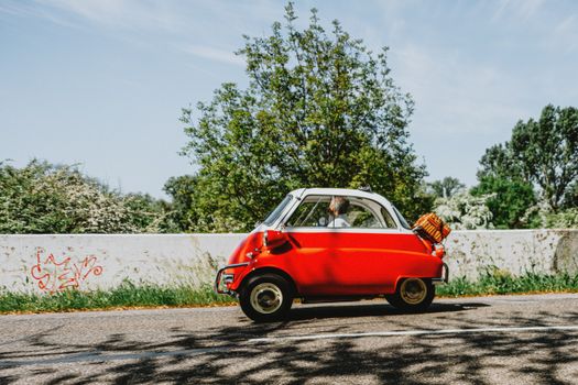 A vintage red microcar cruising on a scenic road with greenery, representing nostalgia and travel.