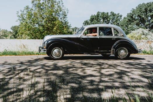 A classic vintage black car driven by an adult on a sunny day, surrounded by greenery.