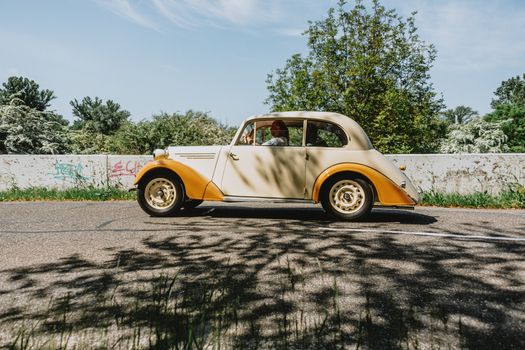 Classic yellow car drives on a sunlit countryside road lined with trees and graffiti.