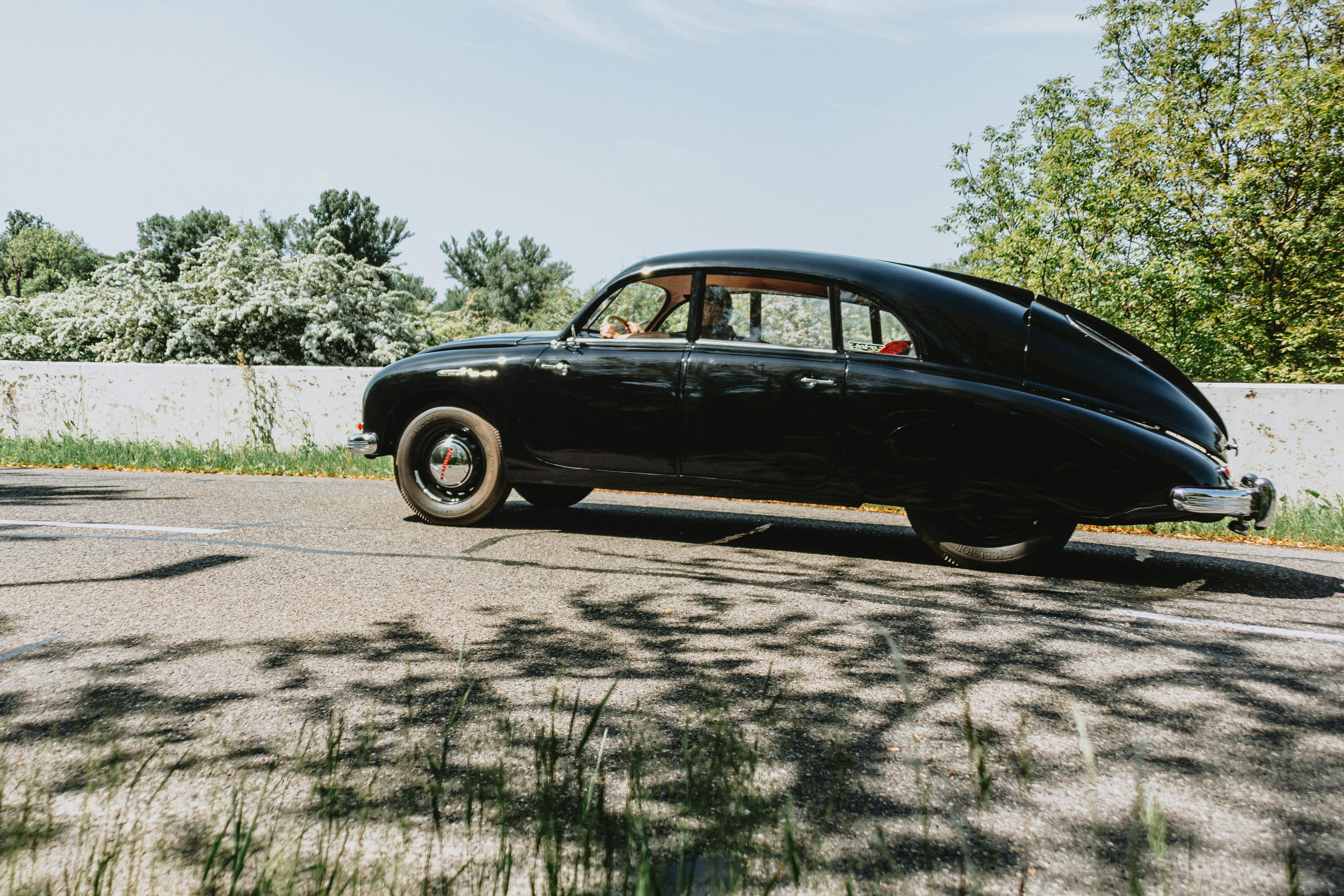 Free Classic black vintage car on a scenic outdoor drive through a sunny, tree-lined landscape. Stock Photo