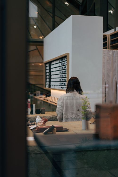 Free A serene interior shot of a Tokyo cafe, capturing an adult female standing at a wooden counter with soft ambient lighting. Stock Photo