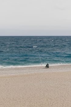 A lone fisherman sits on a beach by the Mediterranean Sea in Antalya, Türkiye, with a fishing pole on a calm day.