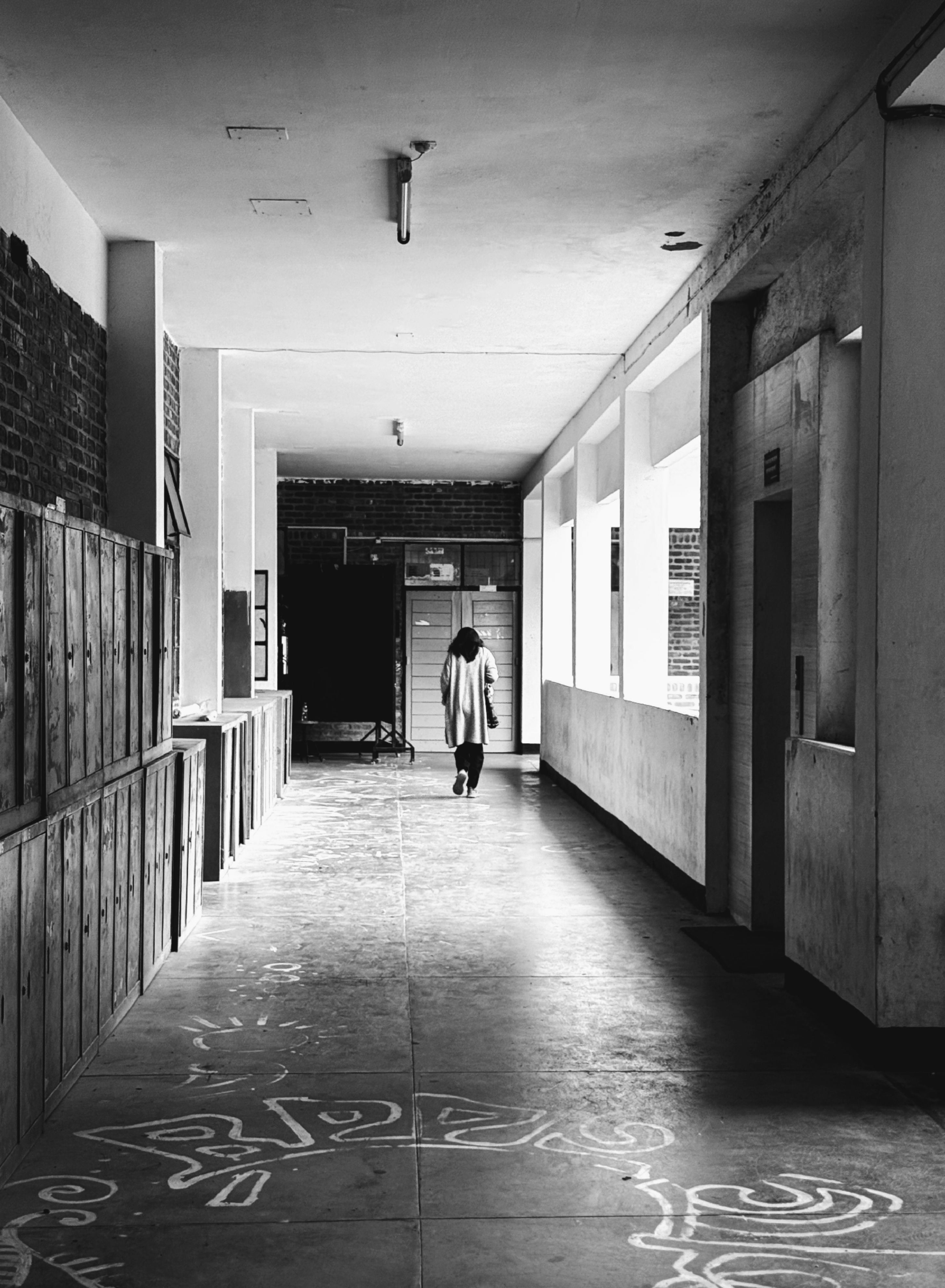 A solitary figure walks down a deserted school corridor in black and white.