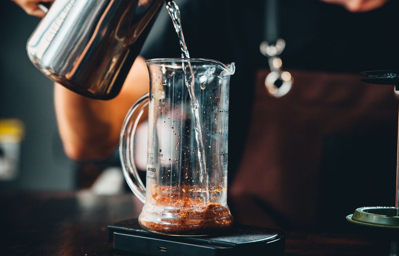 Hands pouring hot water into glass pitcher with coffee grounds for brewing.
