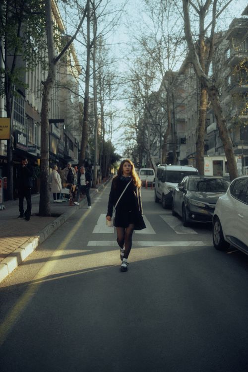 Free A woman in a black outfit strolls down a bustling city street at dusk, surrounded by cars and pedestrians. Stock Photo