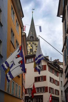 View of Zurich's historic old town street and the St. Peter Church clock tower, flags waving.