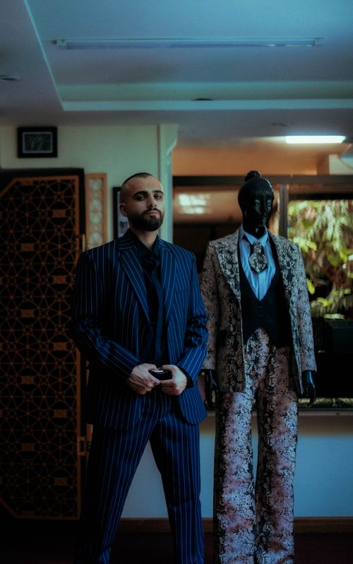 Free Portrait of a stylish man in a striped suit beside a patterned suit on a mannequin in a chic indoor setting. Stock Photo