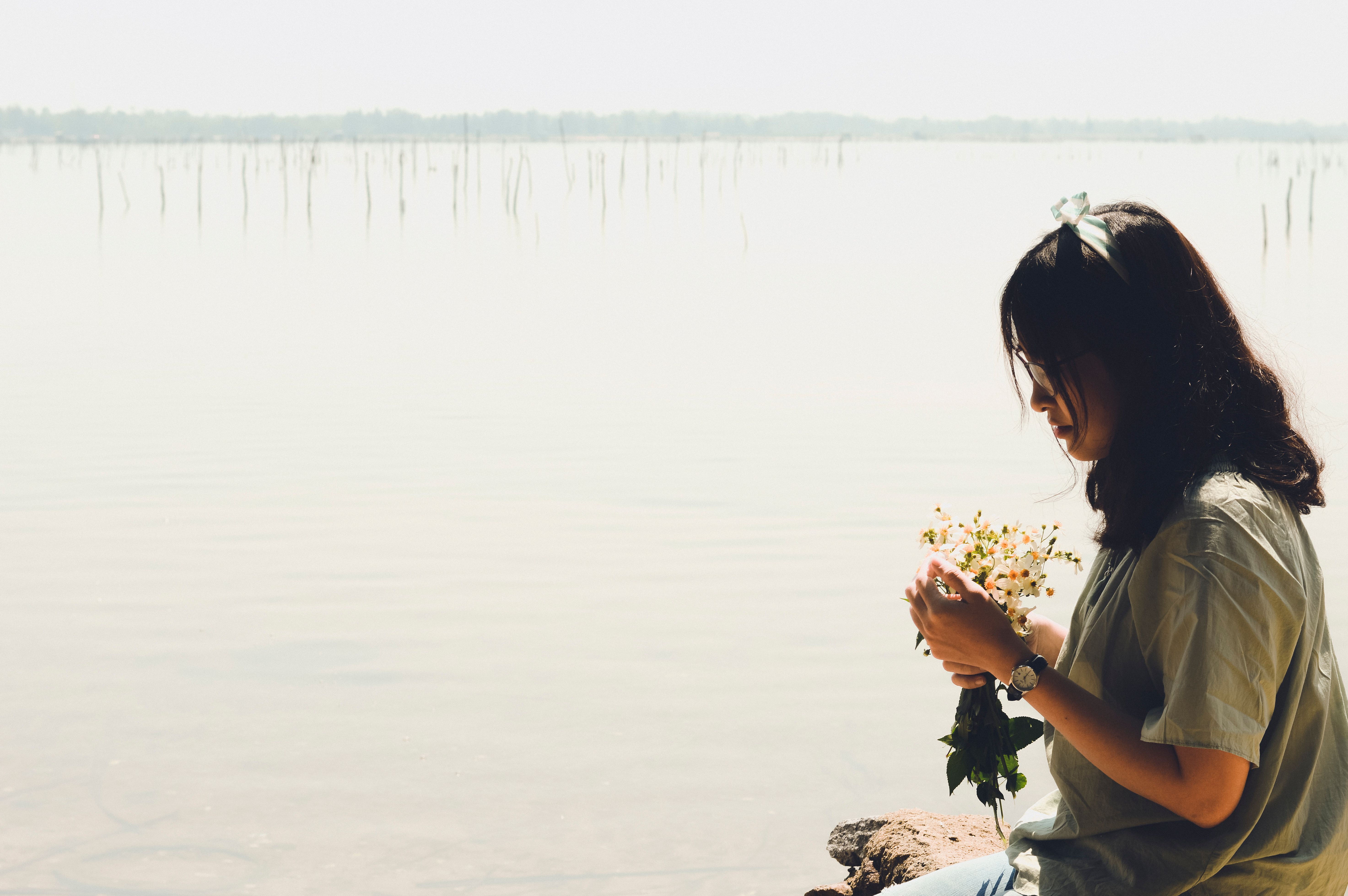 A contemplative young woman holding flowers by a tranquil lakeside in daylight.