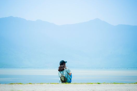 A person sitting alone by a lake, with mountains in the background, under a bright sky.