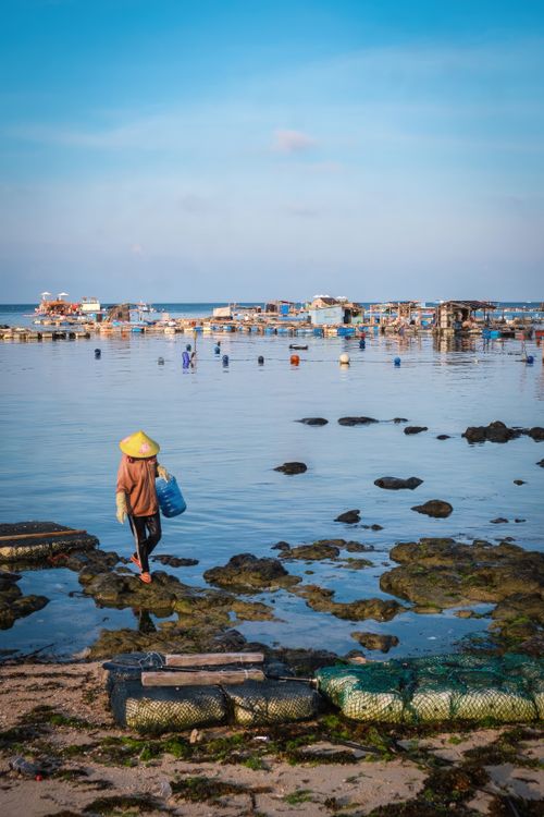 Free A fisherman carries a basket along a rocky shore by a coastal village, with blue skies above. Stock Photo