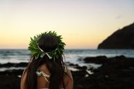 A woman wearing a lei and floral decoration gazes at the ocean during sunset, capturing a tranquil beach moment.