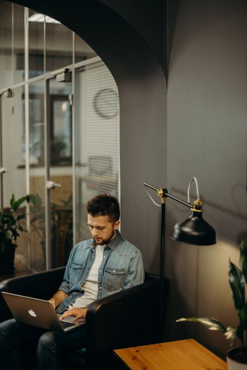 Free Young man sitting in a stylish office working remotely on a laptop with modern decor and natural light. Stock Photo