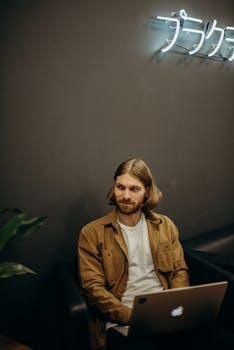 A man with long hair using a laptop in a modern indoor space, illuminated by neon lights.