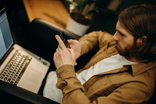 Young man focused on smartphone in a modern workspace with a laptop nearby.