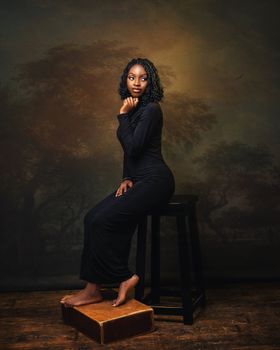 Artistic portrait of a woman in black dress on a stool, warm lighting.
