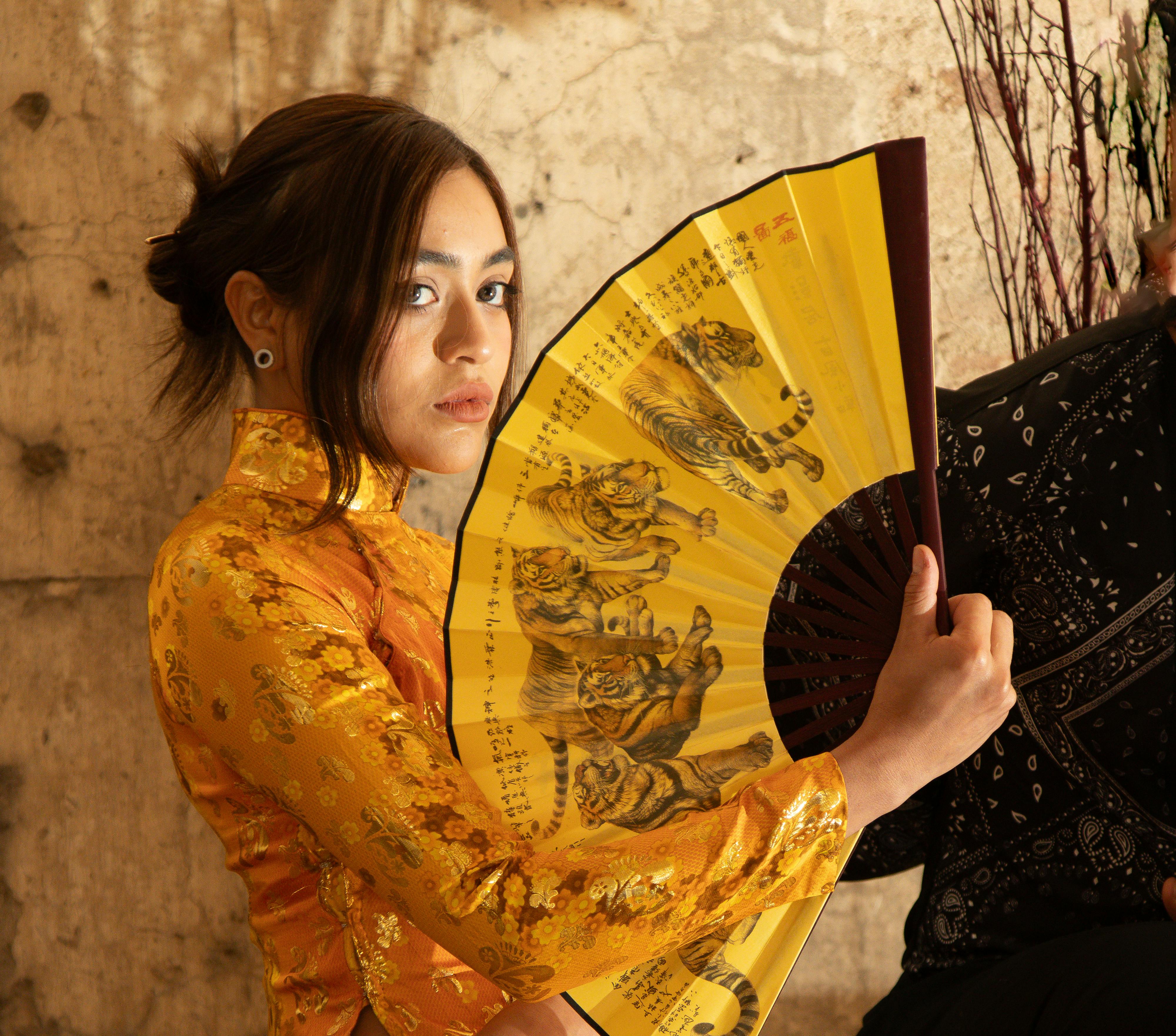 Young woman in a golden traditional dress holding a decorative fan with tiger illustrations indoors.
