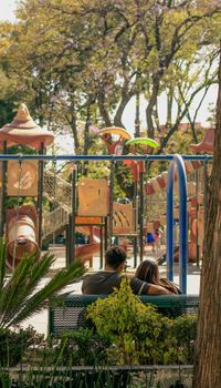 Couple sitting on a bench in a sunny playground at a park in Mexico City, surrounded by trees.