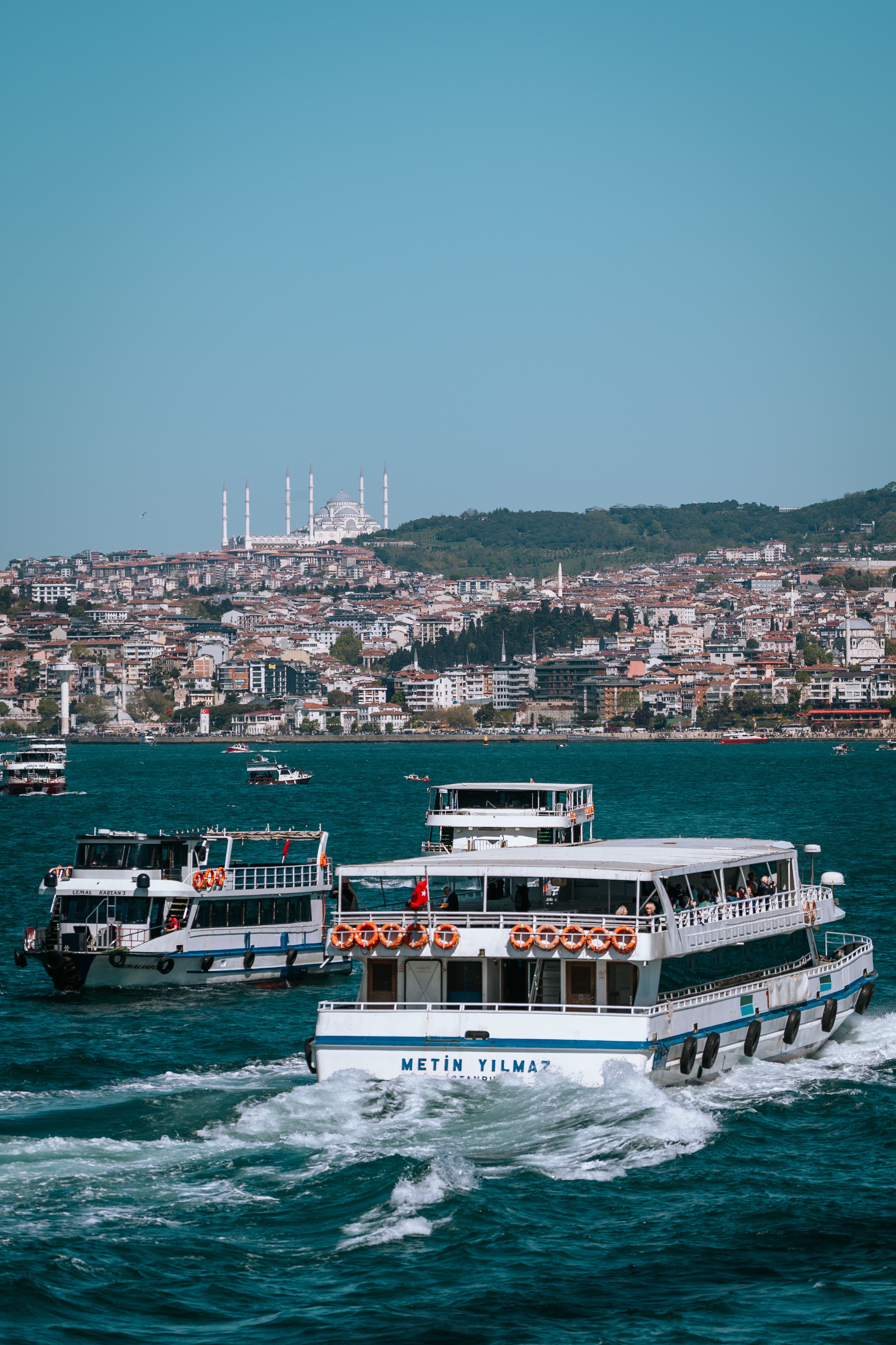 Passenger boats navigating the Bosphorus with a scenic view of Istanbul and the Camlica Mosque in the background.