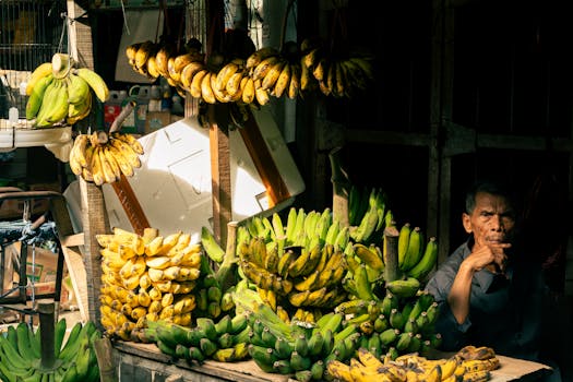 Elderly vendor at a traditional Asian market selling fresh bananas under sunlight.
