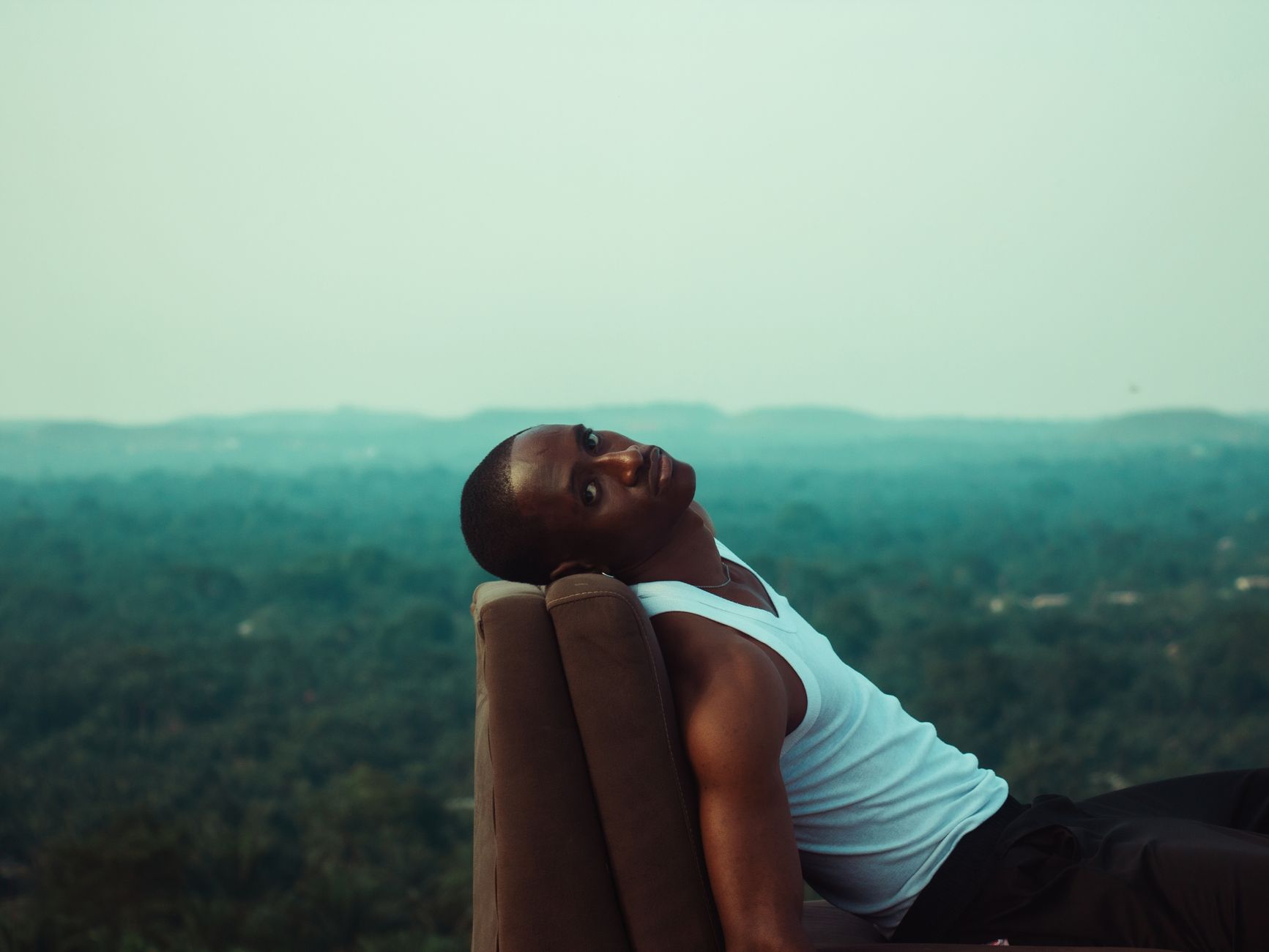 A young man in a white tank top relaxes outdoors, enjoying a scenic view.