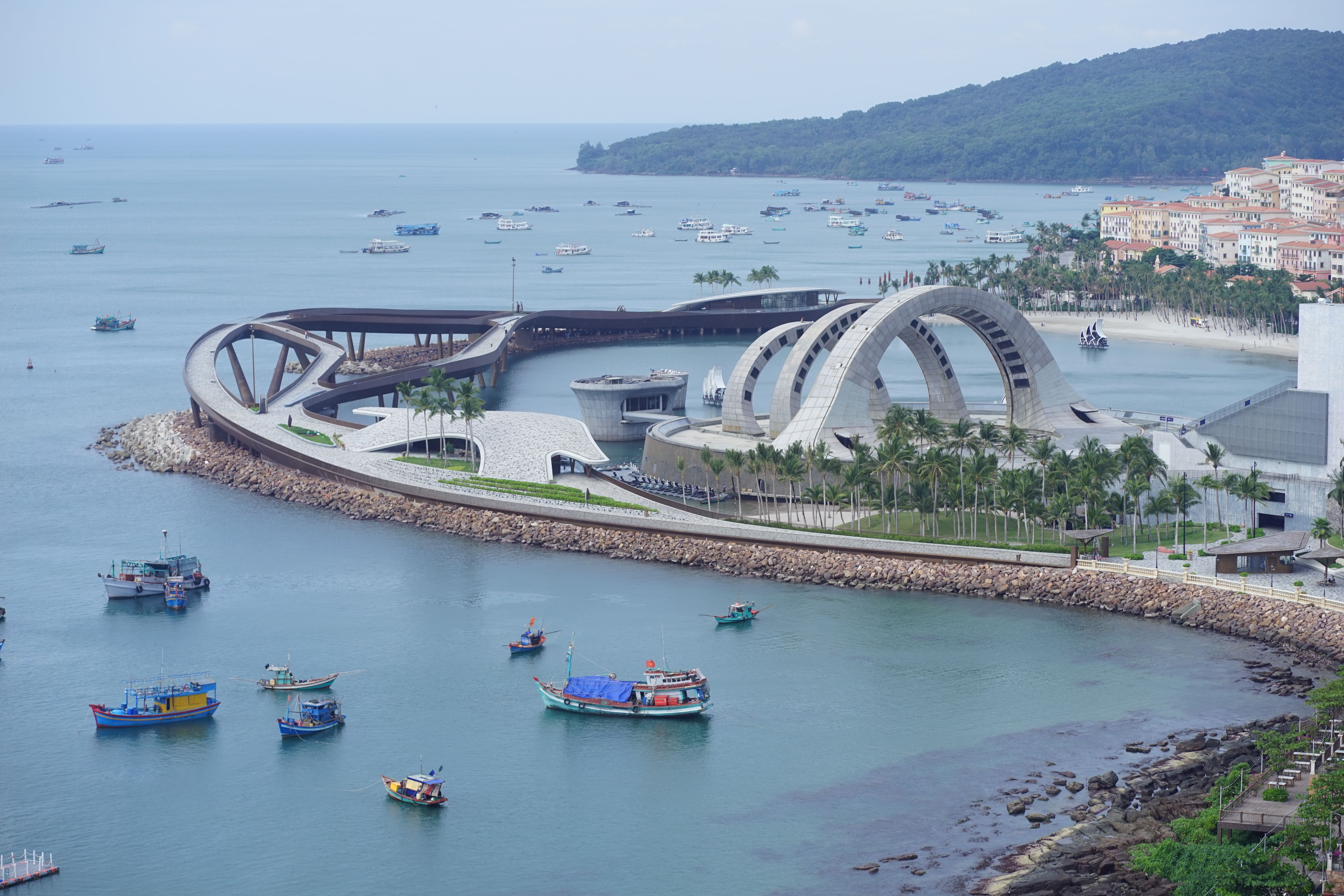 Aerial view of a modern architectural structure by the coast with boats and surrounding landscape.