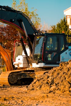 Excavator at a construction site with dirt piles and trees in background.