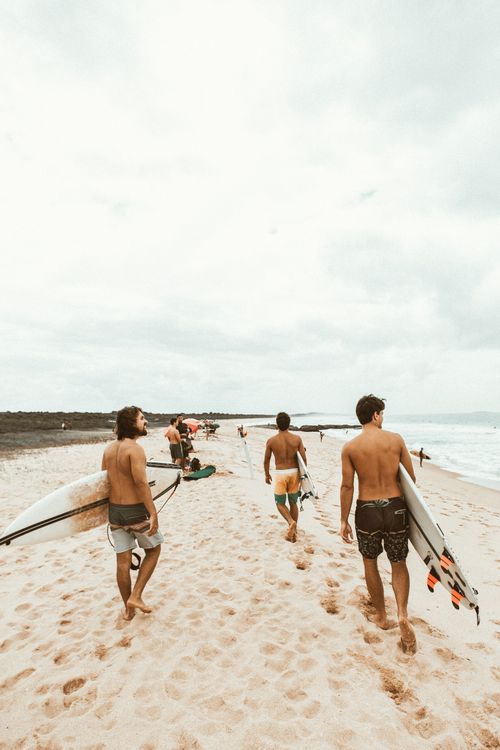 Free Three surfers carry their boards along a sunny beach, ready for water sports adventure. Stock Photo