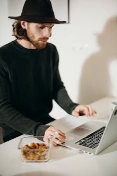 一个戴着帽子的男人在室内用笔记本电脑工作，旁边放着一个食物容器
