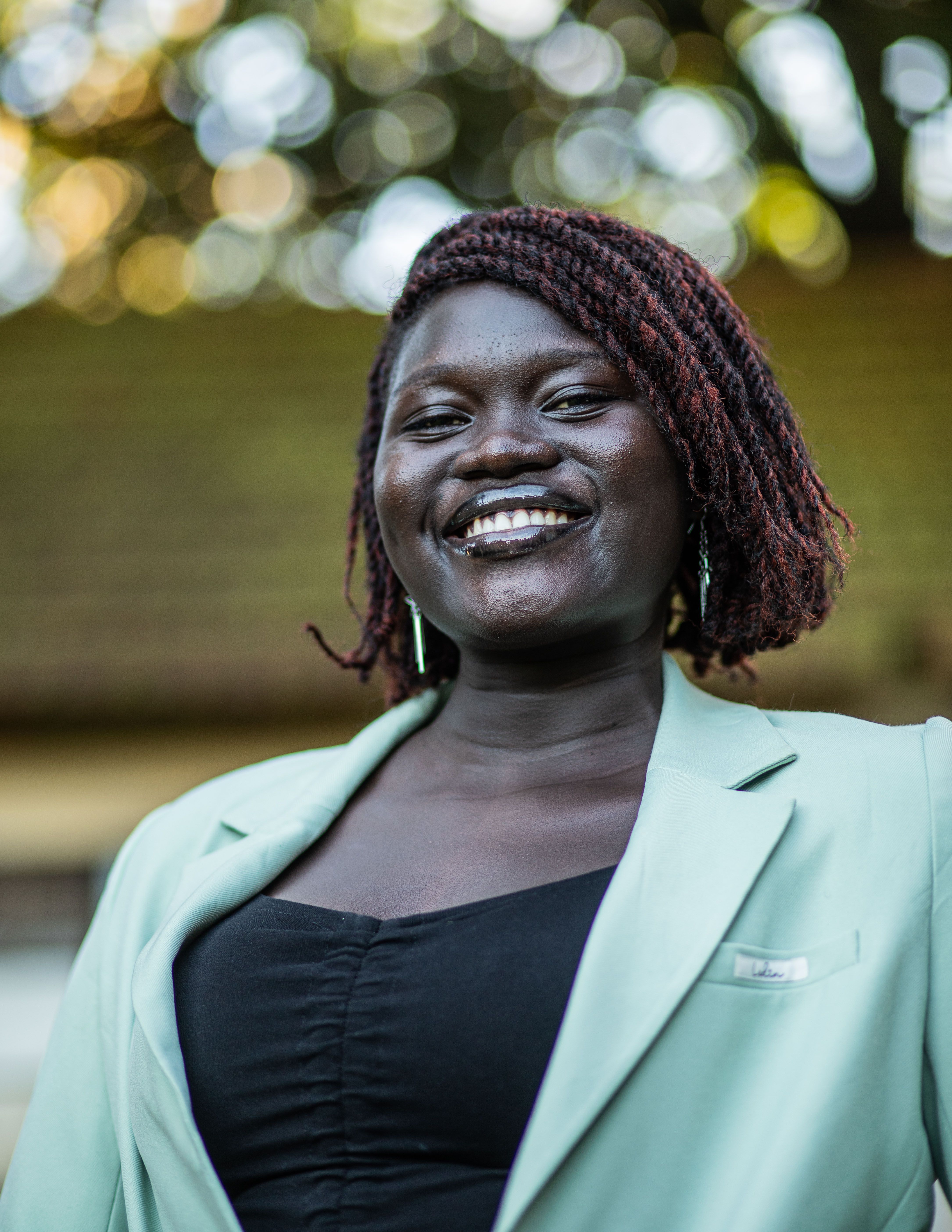 A cheerful African woman in a light green blazer smiling outdoors with a blurred background.